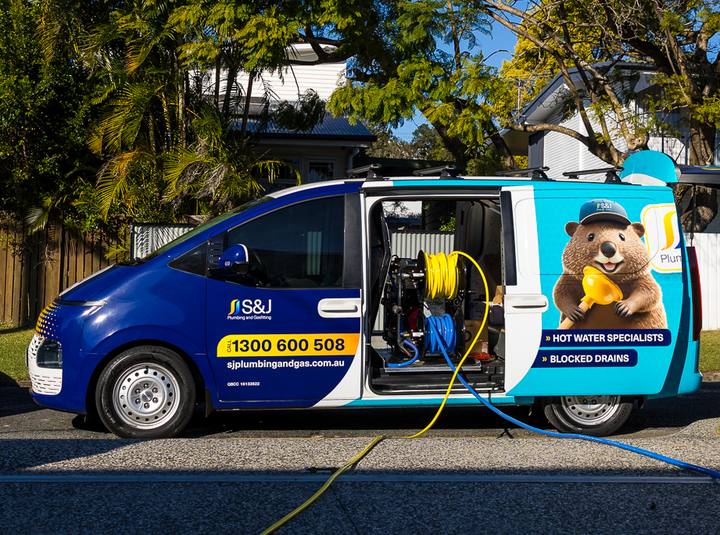 S&J Plumbing van parked on a driveway with its side door open, showing the hose reels and hoses run out for drain clearing