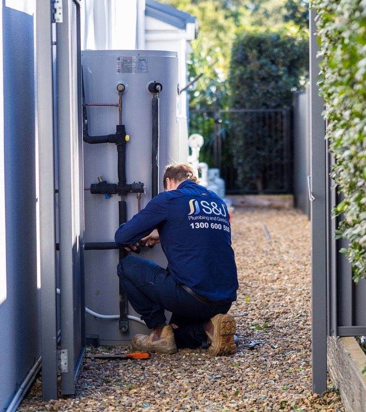 S&J plumber crouched at an outdoor hot water tank in a side passage